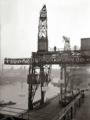 Ein Dreifachkran transportiert vor dem Hafenschuppen 83 im Hamburger Oderhafen drei Kisten an Land. Vor der Laderampe steht ein offener Güterwaggon beladen mit Kisten - auf der Rampe stehen ein Elektrohubwagen und Sackkarren.