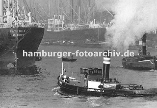 Frachtschiffe liegen im Kaiser-Wilhelm-Hafen - im Vordergrund dampft ein Hafenschlepper durch das Hafenbecken; lks. wird ein Binnenschiff von einem Schlepper geschleppt, dicker Qualm steigt aus dem Schornstein des Arbeitsschiffes. In der Bildmitte