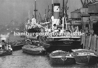 An der Kaianlage des Auguste Victoria Kais im Kaiser-Wilhelm-Hafen liegen Dampffrachter; Kräne löschen die Ladung oder beladen die Schiffe. Auf der Wasserseite der Frachtschiffe haben Schuten fest gemacht - sie werden über das bordeigenes Geschirr