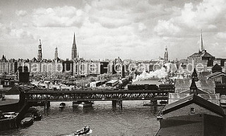 Auf dem Oberhafenkanal fährt eine Barkasse Richtung Oberhafen - auf der Oberhafenbrücke überquert eine qualmende Lokomotive mit Kohlentender und Personenwagen den Kanal.