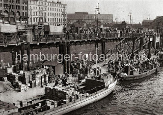 Kähne liegen am Ponton des Deichtormarkts im Oberhafen und werden entladen. Hoch sind die Gemüsekisten und Körbe auf den Lastkähnen und an Land gestapelt. Mit Handkarren und Stechkarren wird die Ladung hinauf zum Markt gebracht.