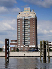 Hochhaus als Touristenhotel am Ufer der Norderelbe. Rechts ragen alte Holzdalben aus dem Wasser.