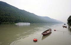 Holzruderboote / Zillen am Ufer der Donau - ein Kreuzfahrtschiff fährt donauabwärts - hohe Berge, Hügel mit Bäumen - Wald am Donaufer.