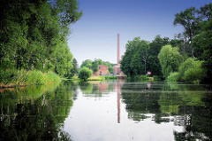 Alte Fabrikgebäude mit hohem Ziegelschornstein von Neu Kaliß spiegeln sich im Wasser des Elde Müritz Wasserwegs. Hohe Bäume und Schilf am Ufer des Wasserwegs.