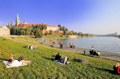 Sonniger Nachmittag in der Grünanlage am Ufer der Weichsel - Wisla. Die Menschen sitzen im Gras oder gehen an der Uferpromenade spazieren. Im Hintergrund der Wawel; Burg - Residenzschloss und Türme der Kathedrale in Krakau / Kraków.
