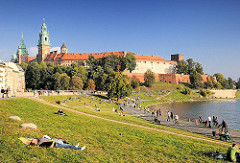 Sonniger Nachmittag in der Grünanlage am Ufer der Weichsel - Wisla. Die Menschen sitzen im Gras oder gehen an der Uferpromenade spazieren. Im Hintergrund der Wawel; Burg - Residenzschloss und Türme der Kathedrale in Krakau / Kraków.