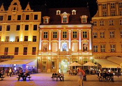 Abend - Nachtaufnahme, beleuchtete Hausfassaden historischer Häuser im polnischen Wraclaw, Breslau. Touristen sitzen im Freien an Tischen unter Markisen oder auf Bänken auf dem Marktplatz.