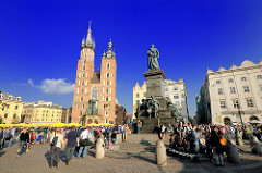Hauptmarktplatz von Krakau  - Wohnhäuser und Marienkirche - Adam-Mickiewicz-Denkmal. Touristen schlendern in der Sonne über den Platz.