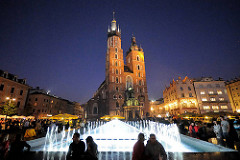 Marktplatz mit Springbrunnen am Abend - Nachaufnahme, beleuchtete Türme des Krakauer Wahrzeichens die Marienkirche.