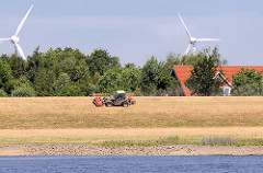 Das Gras des  Elbdeichs wird mit einem Trecker gemäht - im Hintergrund die Rotoren von Windrädern in den Hamburger Vierlanden.