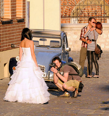Fotoshooting mit Brautkleid / Braut; der Fotograf kniet in kurzen Hosen auf dem Pflaster - küssendes Paar; Strassenszene aus Krakau.
