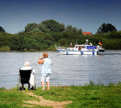 Motorboot auf der Elbe bei Hitzacker - zwei Frauen, eine im Rollstuhl, blicken vom Flussufer  auf das vorbeifahrende Sportboot.