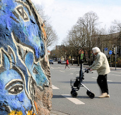 Ausschnitt eines bunt bemalten Felsbrocken - Steinskulpturen der Künstlerin Dagmar Glemme; Frau mit Rollator überquert die Strasse.