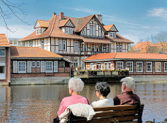 Frühlingstag am Ratsteich, der aufgestauten Ilmenau in Uelzen - Parkbesucher sitzen auf einer Bank in der Sonne am Wasser - auf der gegenüber liegendes Seite des Teichs ein Fachwerkgebäude mit Aussengastronomie an der Ilmenau.