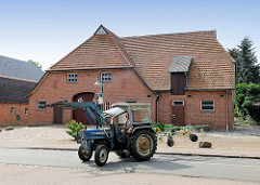 Ein Traktor / Trecker mit angekoppeltem Heuwender auf der Dorfstrasse in Bünningstedt, Gemeinde Ammersbek - im Hintergrund ein landwirtschaftliches Gebäude; Scheune.