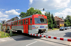Der AKN Zug Richtung Elmshorn überquert die Mühlenstrasse in Barmstedt - die Schranken sind heruntergelassen, Autos warten.