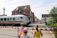Bahnübergang mit heruntergelassener Bahnschranke an der Bahnhofstrasse in Glückstadt - ein Zug fährt vorüber Kinder warten mit ihrem Fahrrad vor der Schranke - im Hintergrund die Große Kremper Strasse.