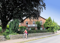 Dicht belaubte Blutbuche / Rotbuche im Vorgarten eines Wohnhauses auf in dem Dorf Bünningstedt, Ammersbek; Steinmauer aus Feldsteinen an der Dorfstrasse - Radfahrerin in Fahrt.