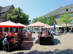 Café / Restaurant in der Fussgängerzone von Ahrensburg / Stormarn; die Gäste sitzen unter Sonnenschirmen im Freien auf dem Strassenpflaster; im Hintergrund die Skulptur Muschelläufer, Künstler Martin Wolke.