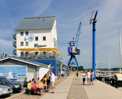 Hafenpromenade in der Stadt Schleswig - ehem. Speicher am Hafenrand und ein alter Hafenkran - Besucher essen Eis; Planung Umbau der Promenade.