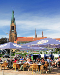 Aussengastronomie am Hafen Schleswigs - Restaurant mit Tischen und Sonnenschirmen  auf der Hafenpromenade - Gäste sitzen in der Sonne - im Hintergrund das Kirchengebäude vom St. Petri Dom.