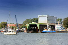 Blick über die Schlei zur Stadt Arnis. Werfthallen am Wasser der Schlei - ein Segelboot fährt unter Motor Richtung Schleswig.