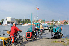 Autofähre / Personenfähre - Seilfähre über die Schlei bei Arnis, Schleswig Holstein. Fahrradfahrer mit Anhänger fahren auf die Seilfähre über die Schlei - im Hintergrund die Stadt Arnis.