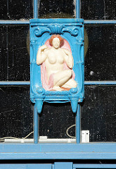 Holzschnitzerei am Fenster vom Eingang U-Bahnstation Schmalenbeck, Gemeinde Grosshansdorf - Vignette nackte Frau in blauem Rahmen.
