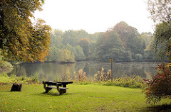 Herbstlich gefärbte Bäume am Mühlenteich in Grosshansdorf; Herbstfarben. Sitzbank / Sitzgruppe aus Holz, Picknickplatz auf einer Wiese am Wasser.