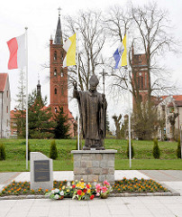 Statue Papst Johannes Paul II. in Pieniężno, Polen.  Im Hintergrund die Kirchtürme der evangelischen und katholischen Kirche und der restaurierte Kupferturm vom Rathaus.