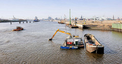 Baggerarbeiten in der Norderelbe - Elbvertiefung; ein Bagger hebt Schlamm vom Flussgrund und füllt den Schlick in eine Schute - im Hintergrund der Kirchenpauerkai in der Hamburger Hafencity.