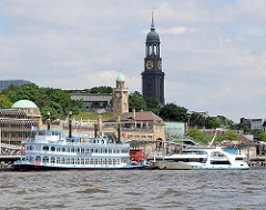 Blick auf die St. Pauli Landungsbrücken - Fahrgastschiffe, Raddampfer liegen für eine Hafenrundfahrt am Ponton - im Hintergrund die Hamburger St. Michaeliskirche / Michel, das Hamburger Wahrzeichen.