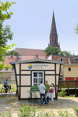 Trompe-l’œil Malerei der Stadtwerke in Pritzwalk - täuschend echte Malerei, Personen stehen an einem Gebäude; im Hintergrund der Kirchturm der St. Nikolaikirche der Stadt in Brandenburg.