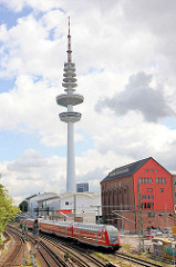 Blick über die Gleise an der Station Hamburg Sternschanze - ein Zug fährt ein; im Hintergrund der Hamburger Fernsehturm, Heinrich Hertz Turm.