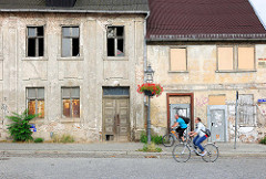 Leerstehende Wohnhäuser, zerbrochene Fensterscheiben / vernagelte Fenster - Blumenschmuck, Ampel mit roten Blumen am Laternenmast - Fahrradfahrer.