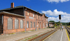 Stillgelegtes Bahnhofsgebäude von Neustadt Glewe; Ziegelgebäude, vernagelte Fenster - Fahrgast auf dem Bahnsteing; blauer Himmel, weisse Wolken.