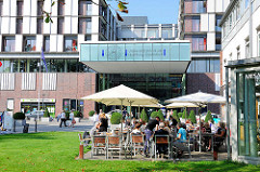 Eingang Neubau Universitätsklinikum Hamburg Eppendorf. Café / Restaurant - Tische + Stühle mit Gästen unter Sonnenschirmen in der Sonne.