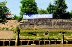 Kaimauer bei Niedrigwasser im Hamburger Hafen - der Bereich bei Hochwasser ist mit Algen bewachsen. Bei Ebbe zeigen sich die Baumstämme, die das Fundament der Kaianlage bilden. Holzdalben sind mit Gräsern bewachsen.