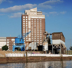 Blick vom Billhafen über die Kaianlagen zum Hotelhochhaus in Hamburg Rothenburgsort. Auf dem Hafenkai steht ein blauer Hafenkran und Verladeeinrichtungen für Schüttgut.