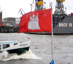 Die Hamburgfahne flattert im Wind - auf der Elbe vor den Landungsbrücken fährt eine Hafenfähre, im Hintergrund Werftanlagen + Dock 10.