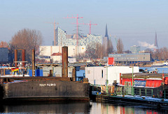 Blick über den Travehafen in Hamburg Steinwerder - Arbeitsschiffe und Schuten liegen am Ponton; am Heck eines grossen Lastkahns ist der Heimathafen Hamburg angebracht - im Hintergrund die Baustelle der Albphilharmonie.