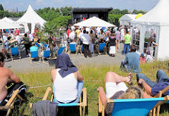 Fest am Spreehafen in Hamburg Wilhelmsburg - Zelte sind aufgestellt; ZuschauerInnen liegen in Liegestühlen auf dem Klütjenfelder Hauptdeich in der Sonne.