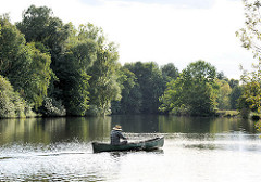 Ein Ruderer mit Strohhut in seinem Ruderboot auf der Dove Elbe - im Hintergrund das Ufer von Hamburg Neuengamme.