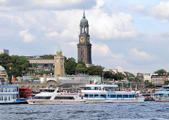 Blick auf die St. Pauli Landungsbrücken - Ausflugsschiffe liegen voll besetzt am Ponton - im Hintergrund der Uhrenturm der Landungsbrücken, dahinter die Jugendherberge und die St. Michaeliskirche.