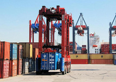 Ein Portalhubwagen transportiert einen Container auf dem Gelände des Terminals EUROGATE - im Hintergrund ein Frachter am Predöhlkai.