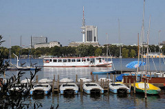 Bootsverleih an der Alster - Tretboote liegen am Bootssteg - ein weisses Schiff der Alsterflotte fährt Richtung Uhlenhorst. Im Hintergrund das Alsterufer von Hamburg Harvestehude, sowie der Fernsehturm und das Radisson Hotel Hamburg