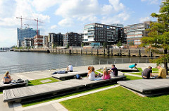 Sommersonne in der Hamburger Hafencity - Hamburg Touristen und Besucher der Hafencity sitzen am Marco Polo Platz - im Hintergrund die Baustelle der Elbphilharmonie und die modernen Wohngebäude am Dalmannkai.