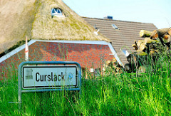 Ortsschild CURSLACK - Holzstapel und Reetdach eines Bauernhauses.