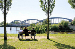 Elbpark von Entenwerder - Grünanlage in Hamburg Rothenbursort - Besucher sitzen zwischen Bäumen auf einer Parkbank und blicken auf die Elbe und die geschwungenen Stahlbögen der Norderelbbrücke.