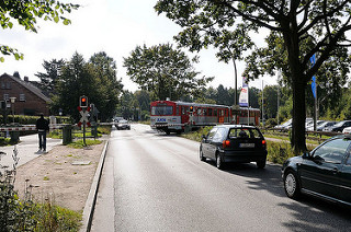 Vorortsbahn - AKN Bahn kreuzt die HolsteinerChaussee. Autos warten vor der Bahnschranke, der Zug fährt in die Haltestelle Burgwedel ein.